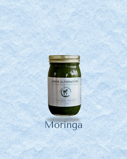 A glass jar of Sheer Alternatives Moringa herbal supplement with a gold lid and white label, centered on a light blue textured background with 'Moringa' text below.