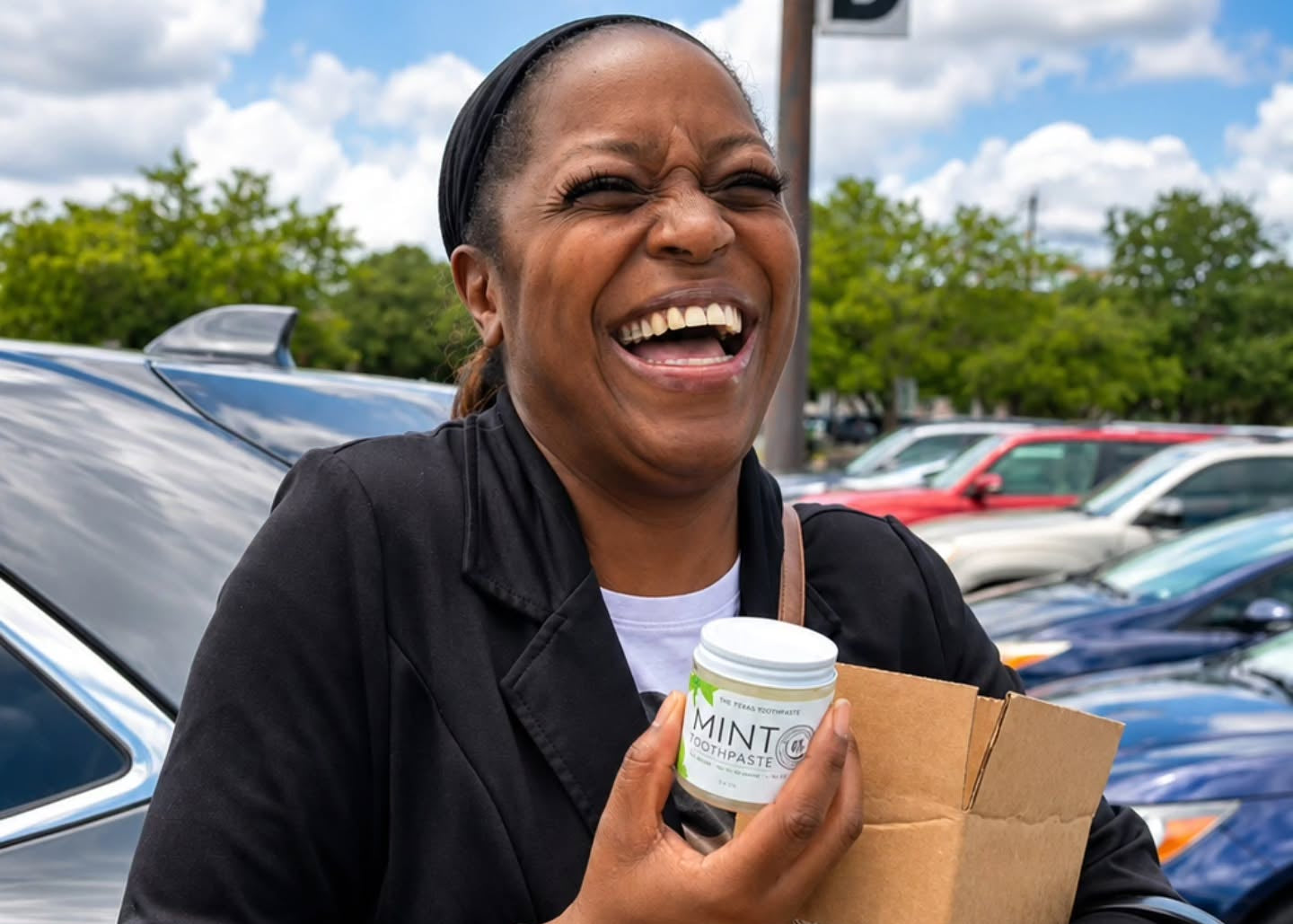 A woman smiling outdoors in a parking lot while holding a Mint 6 mouthwash bottle and a brown cardboard box.